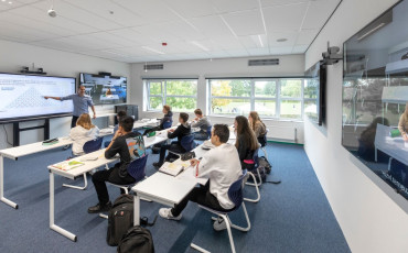 Een onderwijslokaal met een paar studentne die fysiek aanwezig zijn en een student die online onderwijs volgt