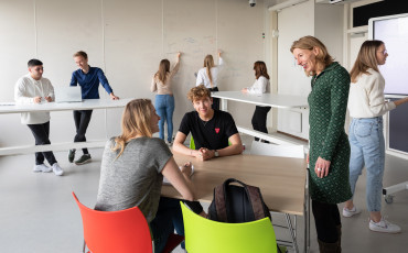 Een docent in gesprek met twee studenten. Studenten op de achtergrond zijn bezig met verschillende leeractiviteiten.