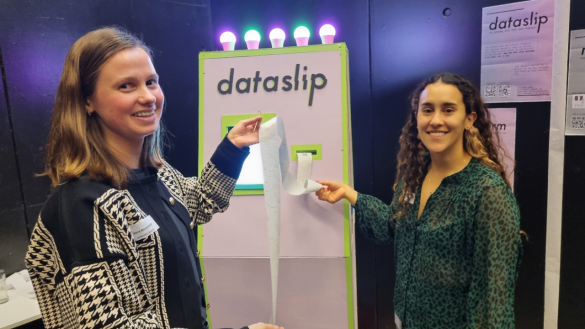 Laura Boeschoten holding her "data slip", a long piece of receipt-looking paper that contains all the data that she has left in different places. On the right is Alejandra Gómez Ortega smiling and holding the end of the receipt. Behind them is the pink "data slip machine" that spit out Laura's data slip.