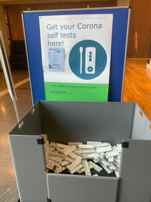 Bak met zelftesten in een hal bij de WUR, poster: "Get your Corona self tests"