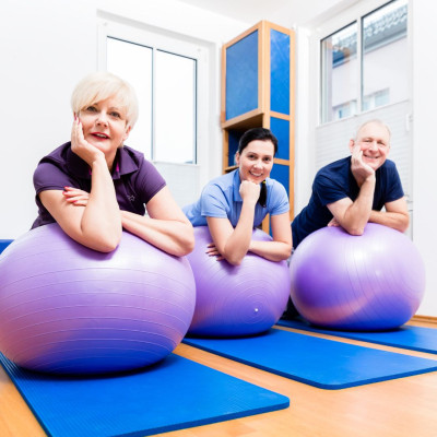 Drie mensen, waaronder twee oudere volwassenen en een jongere persoon, zitten op paarse fitnessballen in een lichte en moderne fitnessruimte. Ze rusten hun kin op hun handen en lijken ontspannen en vrolijk. Op de vloer liggen blauwe yogamatten.