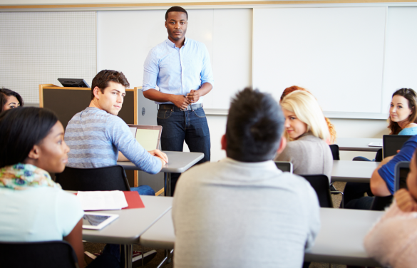 klaslokaal met een diverse groep studenten die aan tafels zitten en aandachtig luisteren naar een staande docent. De docent, een jonge man in een blauw overhemd, kijkt richting de studenten terwijl hij lijkt te spreken. De studenten tonen verschillende reacties, zoals luisteren, aantekeningen maken en elkaar aankijken. De ruimte heeft een groot whiteboard op de achtergrond