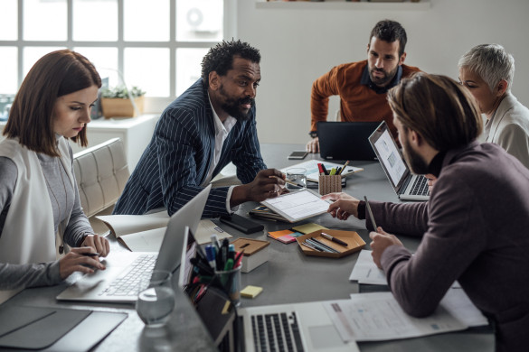 vijf personen overleggen met elkaar aan tafel met laptops en notitieblokken 